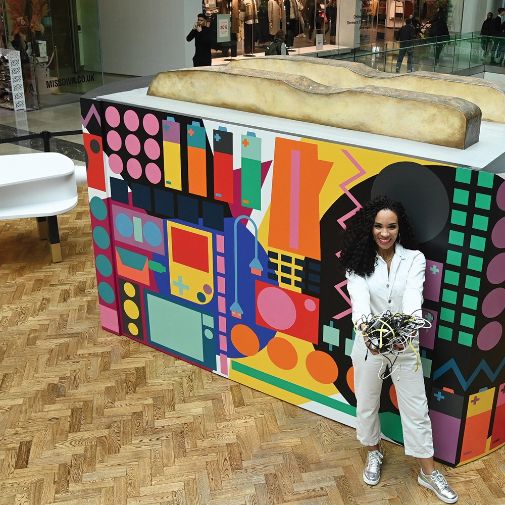 dark skinned presenter Michelle Ackerley in front of giant multi-coloured patterned toaster in a shopping centre, holding a handful of cables