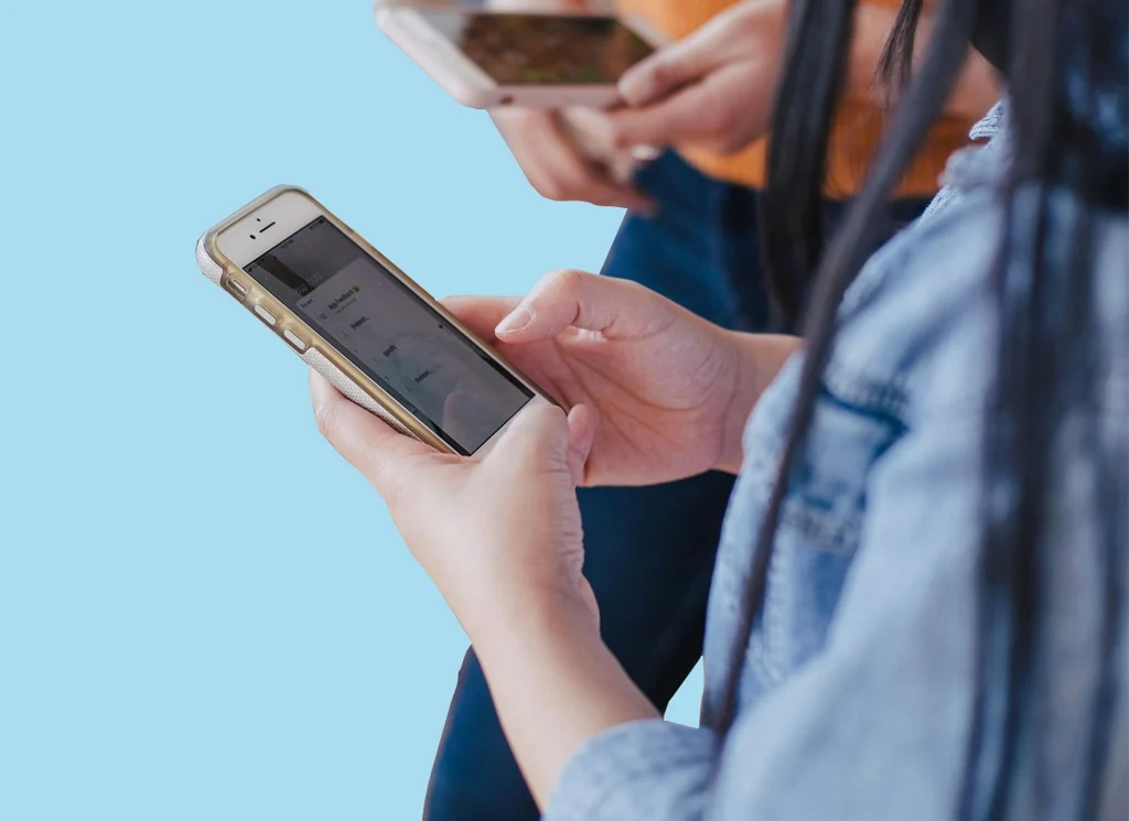 Medium light skinned young person using a smart phone, with someone else using a phone in the background.
