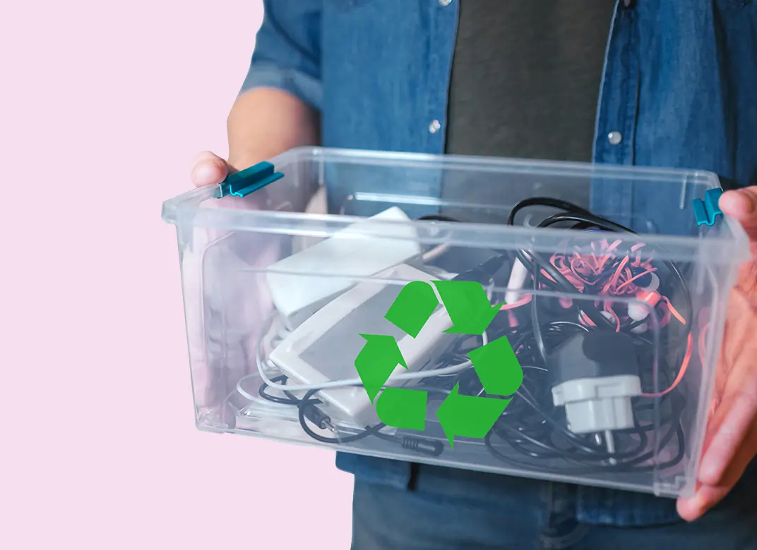 A light skinned man holding a see through plastic box full of electrical goods with a green recycle logo on the front against a pink background.