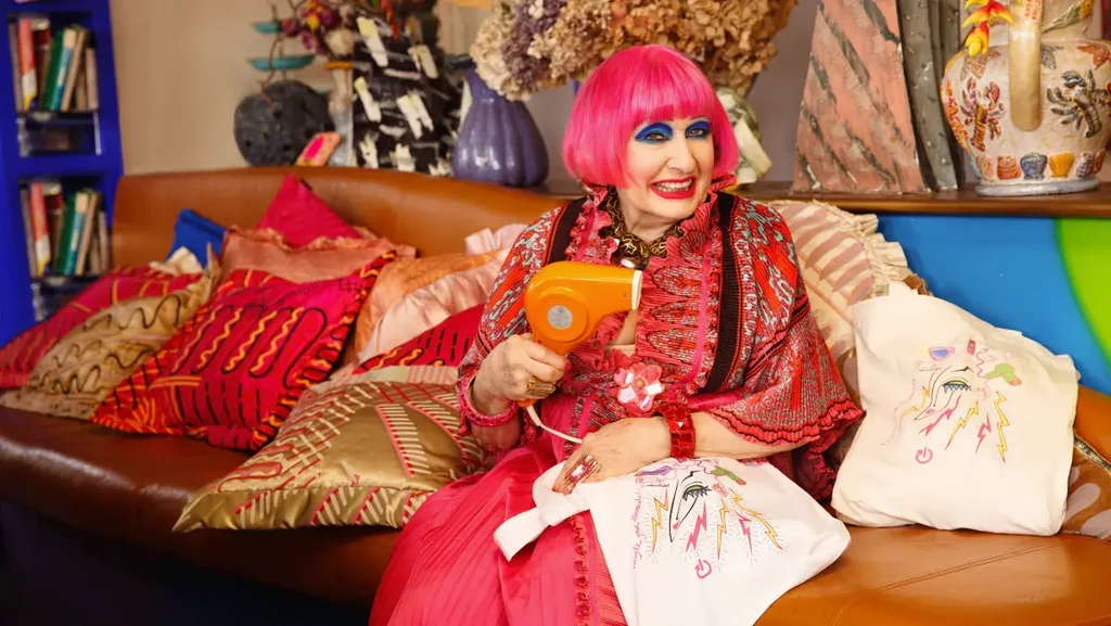 Zandra Rhodes spring clean tote 1200x676px Light skinned fashion designer Zandra Rhodes wearing pink with pink hair holding electrical tote bag she designed and a hairdryer, sitting on a sofa with bright cushions